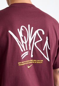 Pessoa vestindo uma camiseta Nike cor vinho com grande texto branco em estilo graffiti "Nike" e uma pequena declaração de marca amarela e logótipo do swoosh nas costas.