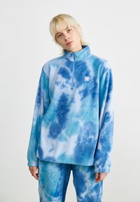 Blå tie-dye fleecegenser med halv glidelås, med en myk tekstur og et lite logo-patch på venstre bryst. Lange ermer.