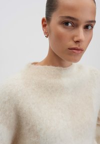 Cream-colored sweater with a soft, fuzzy texture, round neckline, and minimal seams. Model wears a silver stud earring for detail.