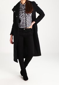 Cappotto lungo nero con bottoni doppi, camicia a quadri corta, jeans skinny neri e stivali neri alla caviglia. Tessuti dal finish liscio.