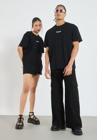 Černé oversized trička s bílým textem, spárovaná s cargo kalhotami a platformovými sandály. Hladká bavlněná textura a uvolněný střih.