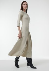 Vestido midi en beis claro con mangas de tres cuartos, con un cierre frontal y diseño plisado, combinado con botas de tacón alto negras.