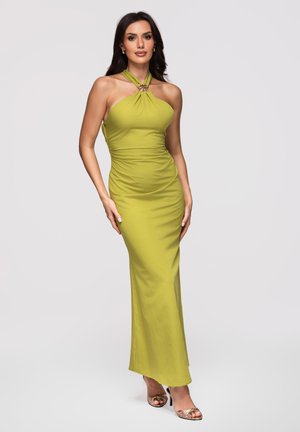Femme debout portant une robe dos-nu vert citron avec une broche décorative au niveau de l'encolure et des talons métalliques à bout ouvert.