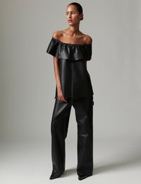 Μαύρο δερμάτινο τοπ με έξω τους ώμους και λεπτομέρεια με βολάν, συνδυασμένο με μαύρο δερμάτινο παντελόνι με φαρδιά μπατζάκια, διακοσμημένο με λευκό ραφή και λεπτομέρειες με καρφιά.