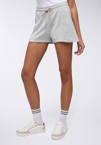 Pantaloni corti in morbido tessuto grigio chiaro con cintura elastica, cordino e tasche laterali, abbinati a scarpe da ginnastica bianche e calzini alla caviglia a righe.