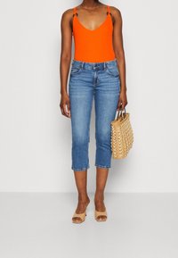 Orange ärmlös bodysuit, blå korta jeans, beiga klacksandaler och vävd tan handväska med axelremmar och framfickor.