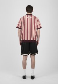 Homme de dos portant une chemise à manches courtes rayée rose et bordeaux, un short noir avec une bordure beige, des chaussettes blanches et des chaussures noires.