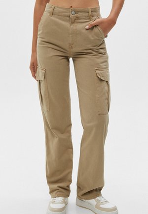 Femme portant un pantalon cargo beige avec des poches à rabat sur les côtés et des baskets blanches, avec une main dans la poche.