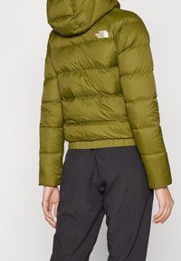 Olivově zelená naducaná bunda s kapucí, která má prošívané části a elastický spodní lem. Logo na levé části hrudi; doplněná černými kalhotami.