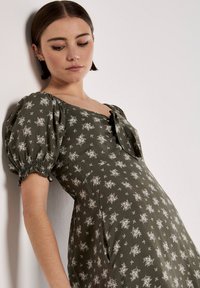Robe vert olive avec de courtes manches bouffantes, un détail de nœud, et un motif floral blanc, fabriquée à partir d'un tissu doux avec un léger éclat.
