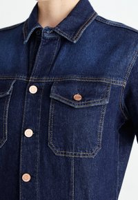 Veste en denim foncé avec une fermeture à boutons sur le devant, deux poches poitrine, des surpiqûres orange contrastantes et des accents de quincaillerie en bronze.