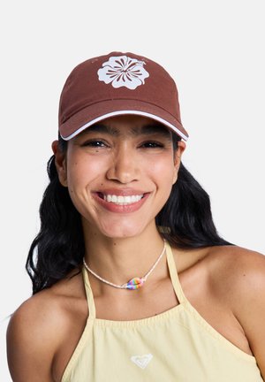 Lächende Frau mit langen dunklen Haaren, die eine braune Mütze mit weißer Blume, ein gelbes Neckholdertop und eine bunte Muschel-Halskette vor weißem Hintergrund trägt.