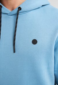Sudadera azul claro con capucha triangular, tejido texturizado, cordones negros y un pequeño logo redondo negro en el pecho.