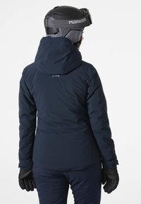 Marineblaue Skijacke mit figurschmeichelndem Design, ausgestattet mit einer abnehmbaren Kapuze und schwarzen Handschuhen. Glatte Textur und minimales Branding auf dem Rücken.