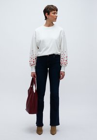 Λευκό φούτερ με κόκκινα σχέδια καρδιών στα μανίκια, συνδυασμένο με σκούρο μπλε τζιν. Το μοντέλο κρατάει κόκκινη τσάντα tote και φοράει μπεζ μπότες με τακούνι.