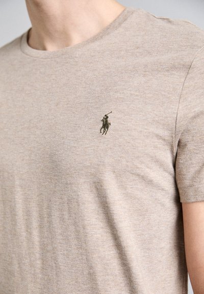 Bežinė marškinė su minkšta, lengva medžiaga; krūtinėje yra mažas tamsiai žalias siuvinėtas logotipas; apvalus kaklas ir trumpi rankovės.