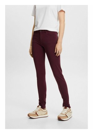 Personne portant un pantalon violet foncé ajusté, un t-shirt blanc et des baskets beige, debout sur un fond clair uni.