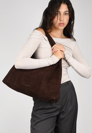 Bolso de hombro de ante marrón con una correa ancha, que presenta una textura suave y una forma amplia, sostenido contra un fondo neutro.