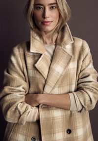 Cappotto a quadri beige con un tessuto morbido e testurizzato. Presenta un ampio colletto, una chiusura doppiopetto e due bottoni. Indossato sopra un maglione leggero.