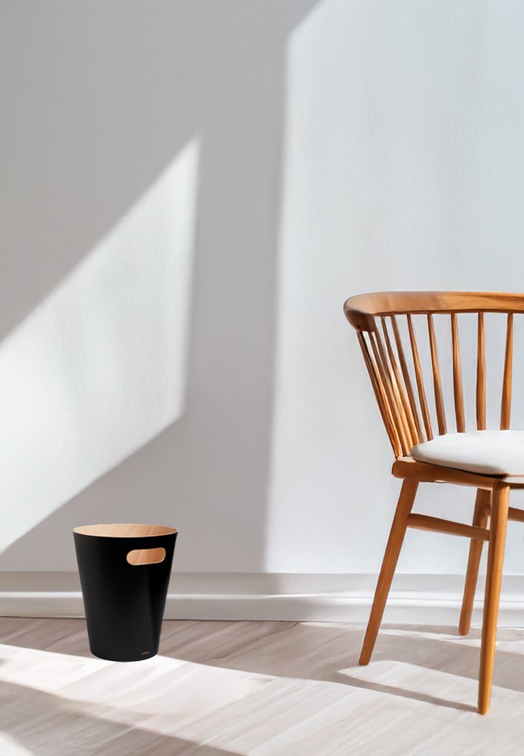 Schwarzer Metall-Abfallbehälter in runder Form mit ausgeschnittenen Griffen, ausgestattet mit einem Innenleben aus natürlichem Holz. Positioniert in der Nähe eines Holzstuhls.