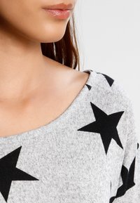 Haut en tricot gris avec un col large et des motifs d'étoiles noires. Texture douce avec une coupe décontractée, mettant en valeur une partie de la zone de la clavicule.