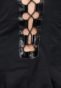 Μαύρο ένδυμα με γυαλιστερή δερμάτινη επένδυση που διαθέτει διασταυρωμένα κορδόνια, αποκαλύπτοντας το δέρμα από κάτω σε κάθετο οβάλ μοτίβο.