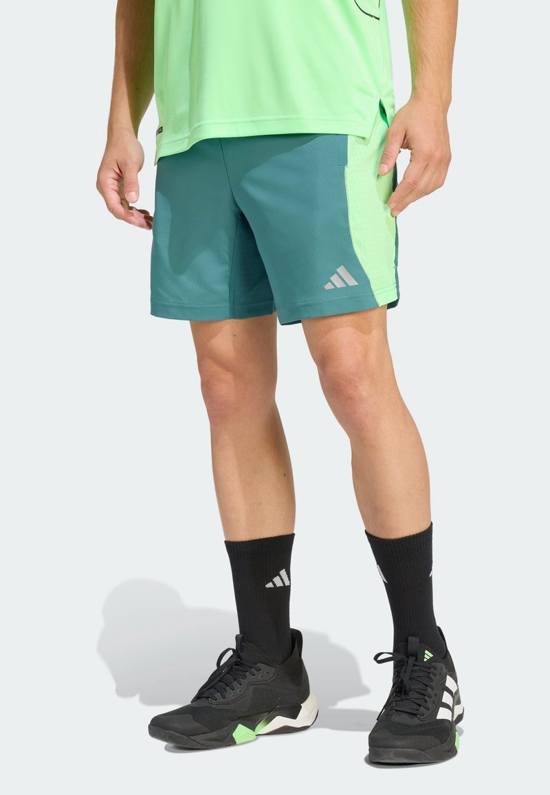 Personne portant un short de sport bleu sarcelle et vert clair, des chaussettes Adidas noires et des chaussures de sport noires Adidas, debout devant un fond uni.