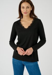 Zwarte langslaper top met een V-hals en kantafwerking. De stof is zacht en heeft een gladde textuur. Gecombineerd met blauwe jeans.