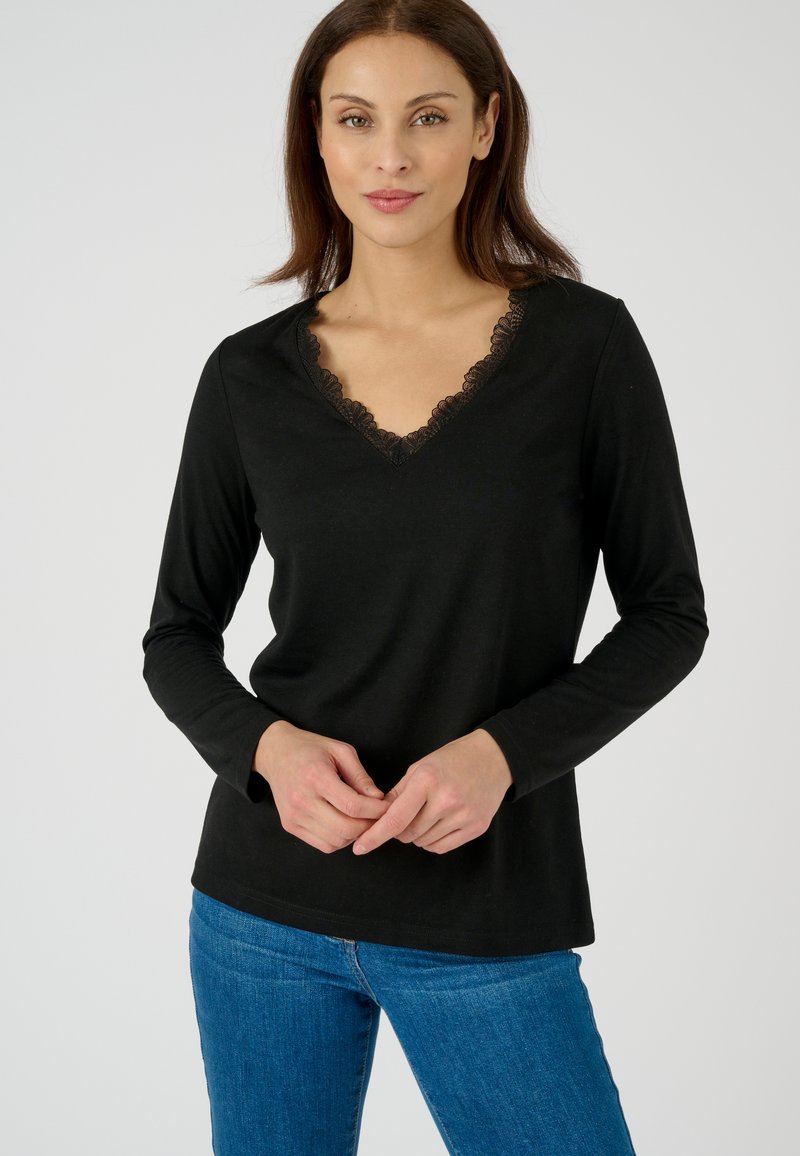 Zwarte langslaper top met een V-hals en kantafwerking. De stof is zacht en heeft een gladde textuur. Gecombineerd met blauwe jeans.