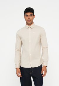 Beige langermet skjorte med krage og knapper, med en liten logo på brystet. Glatt stoff, tilpasset passform med rett falder.