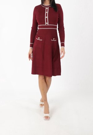 Woman wearing a knee-length long-sleeve burgundy dress with white trim and gold buttons, paired with white open-toe heels.