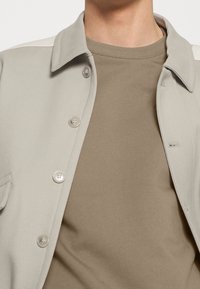 Chaqueta de botón gris claro con botones plateados y un bolsillo en el pecho, superpuesta sobre una camiseta taupe. Tejido suave, diseño moderno.