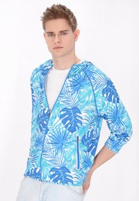 Un tânăr îmbrăcat cu un hanorac cu fermoar, cu imprimeu de frunze tropicale în nuanțe de albastru și turcoaz, peste o cămașă albă, purtând blugi din denim deschis, stând în fața unui fundal alb.