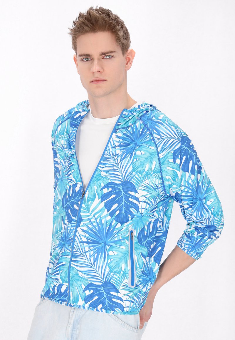 Nuori mies, jolla on päällään sininen ja turkoosi trooppisen lehtien kuvioitu vetoketjullinen huppari valkoisen paidan päällä, kevyet farkut yllä, seisoo valkoista taustaa vasten.
