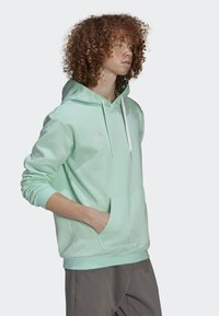 Muntgroene hoodie met een voorzak, een verstelbare trekkoordcapuchon en een wit Adidas-logo. De stof lijkt zacht en comfortabel te zijn.