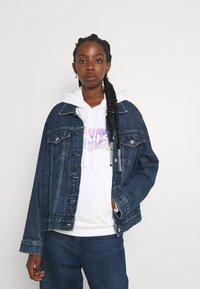 Denimjakke i mørkeblått, med en løs passform, brystlommer og knappelukking over en hvit hettegenser med et fargerikt grafisk trykk.