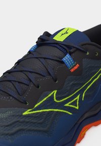 Σκούρο μπλε αθλητικό παπούτσι με νεον πράσινο λογότυπο και λεπτομέρειες, μπλε κορδόνια και πορτοκαλί λεπτομέρεια στη σόλα, σε κοντινή όψη από το πλάι.