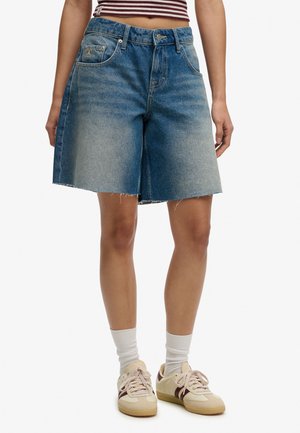 Donna che indossa pantaloncini di jeans blu sbiaditi fino al ginocchio con orlo grezzo, abbinati a calzini bianchi, sneakers crema e un top corto a righe.