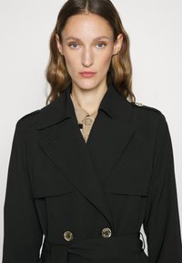 Svart trenchcoat med dobbeltknappet design, gullknapper, bred krage og frontlommeposer. Glatt tekstur, belte i midjen.