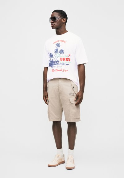 Mann trägt übergroßes weißes T-Shirt mit Grafik, beigefarbene Cargoshorts, weiße Socken, cremefarbene Sneakers und große transparente Sonnenbrille, steht vor schlichtem Hintergrund.