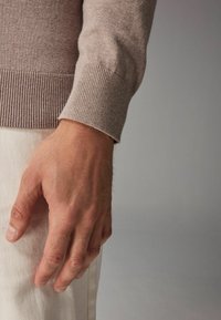 Világosbarna pulóver bordázott mandzsettákkal, fehér nadrág felett viselve. Közeli kép egy kézről, amely a szöveten pihen, kiemelve a textúrát és a színkontrasztot.