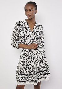 Schwarz-weiß gemustertes Kleid mit V-Ausschnitt, langen Ärmeln und gestuftem Design, das an dem Saum mit einem geometrischen Besatz versehen ist. Weiche Stofftextur.
