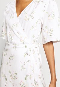 Robe portefeuille en tissu blanc léger avec un motif floral aux accents roses et verts. Comprend des manches courtes et un détail à la taille avec un lien.