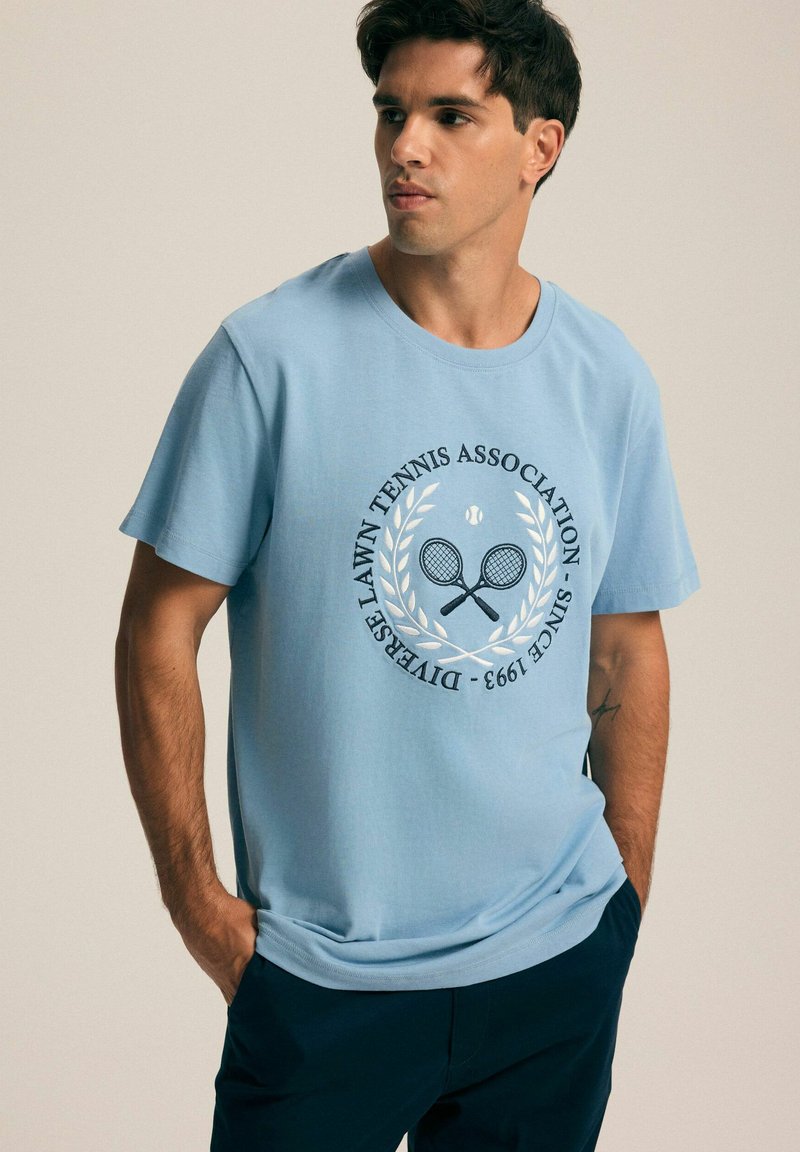 Jasnoniebieska bawełniana koszulka z okrągłym nadrukiem przedstawiającym skrzyżowane rakiety tenisowe i liście laurowe, tekst: "DIVERS LAWN TENNIS ASSOCIATION SINCE 1993."