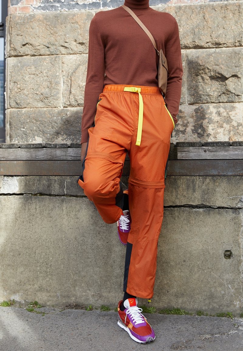 Personne portant un col roulé marron, un pantalon orange avec un cordon jaune, des baskets violettes et rouges, et un sac bandoulière beige, debout à l'extérieur.