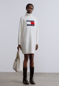 Fehér, túlméretezett pulóverruha garbónyakkal, tengerészkék, piros és fehér márkajelzéssel. Barna, térdig érő csizmával és világos színű táskával.