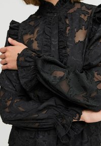 Schwarz durchscheinende Bluse mit strukturiertem Blumenmuster, Rüschenkragen und -manschetten. Verfügt über einen Knopfverschluss und geschichteten Stoff für Tiefe.
