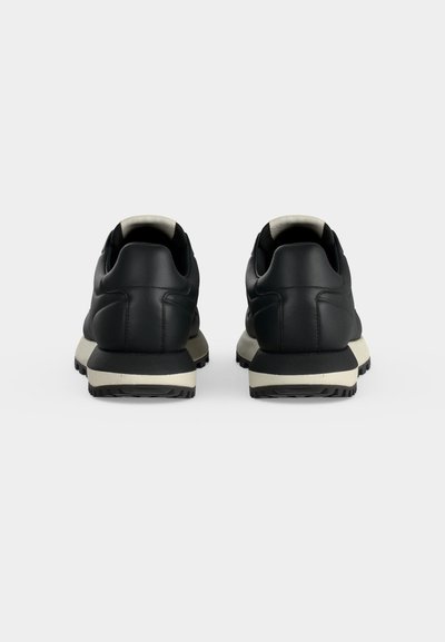 Paire de baskets noires avec semelles intermédiaires blanches, vues de dos sur un fond gris uni.