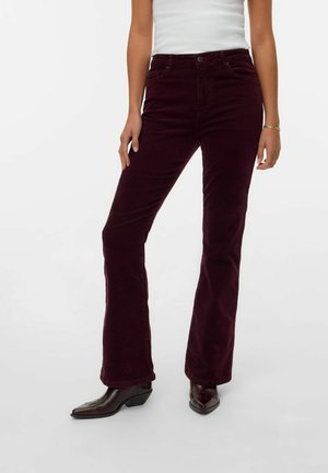 Vrouw in donker bordeauxrode corduroy broek met een witte mouwloze top en donkerbruine spitse enkellaarzen, staande tegen een witte achtergrond.