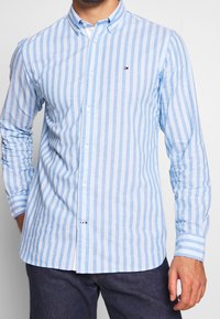 Light blue striped shirt made of cotton. Features a button-down collar, long sleeves, and small logo on the chest. Tailored fit.